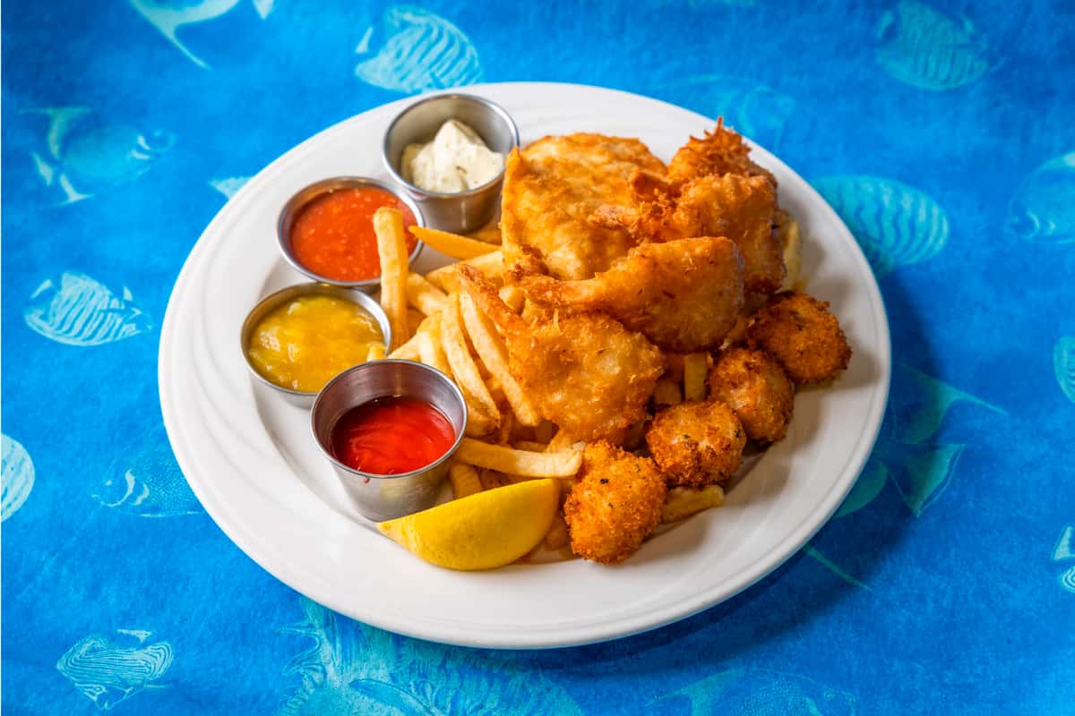 Crispy fried fish fillets and breaded shrimp served with golden fries, lemon wedge, tartar sauce, ketchup, cocktail sauce, and honey mustard on a white plate over a blue ocean-themed tablecloth.