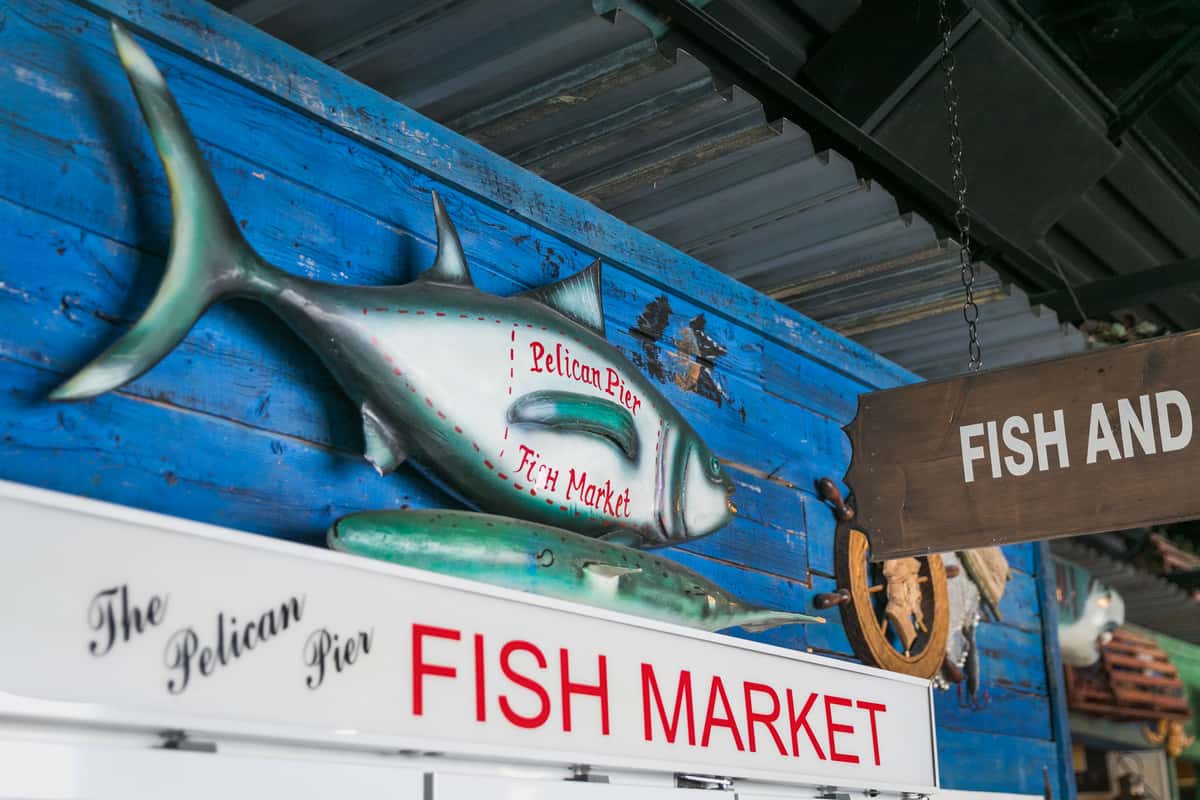 "Decorative fish sign reading 'Pelican Pier Fish Market' mounted on a rustic blue wooden wall above the seafood display area in a nautical-themed restaurant.