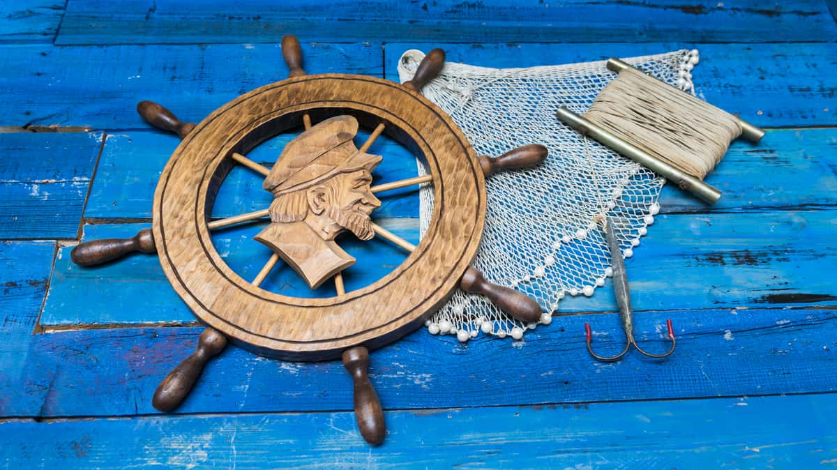 "Rustic nautical decor featuring a wooden ship wheel with a carved sailor's profile, alongside fishing net, coiled twine, and a large fishing hook on a weathered blue wooden background.