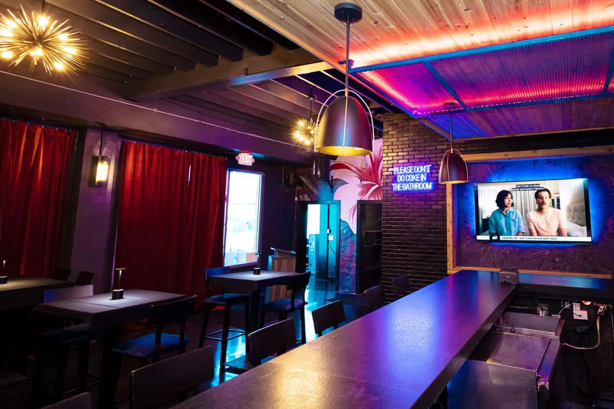 Modern bar interior with moody lighting, red curtains, a neon sign, TV screen, pendant lights, and sleek high-top tables.