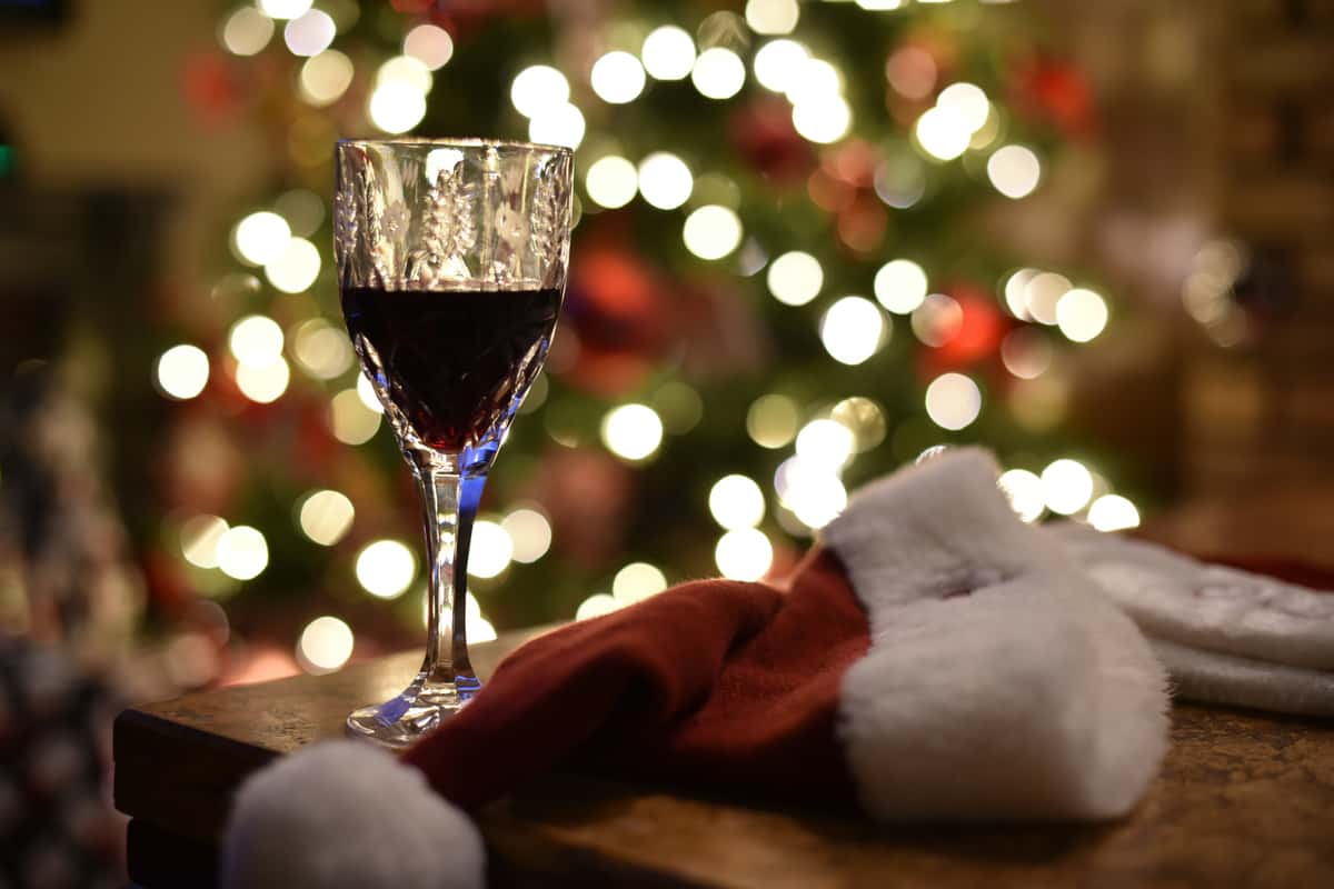 Crystal glass of red wine beside a Santa hat on a wooden table, with blurred Christmas lights and tree in the background.