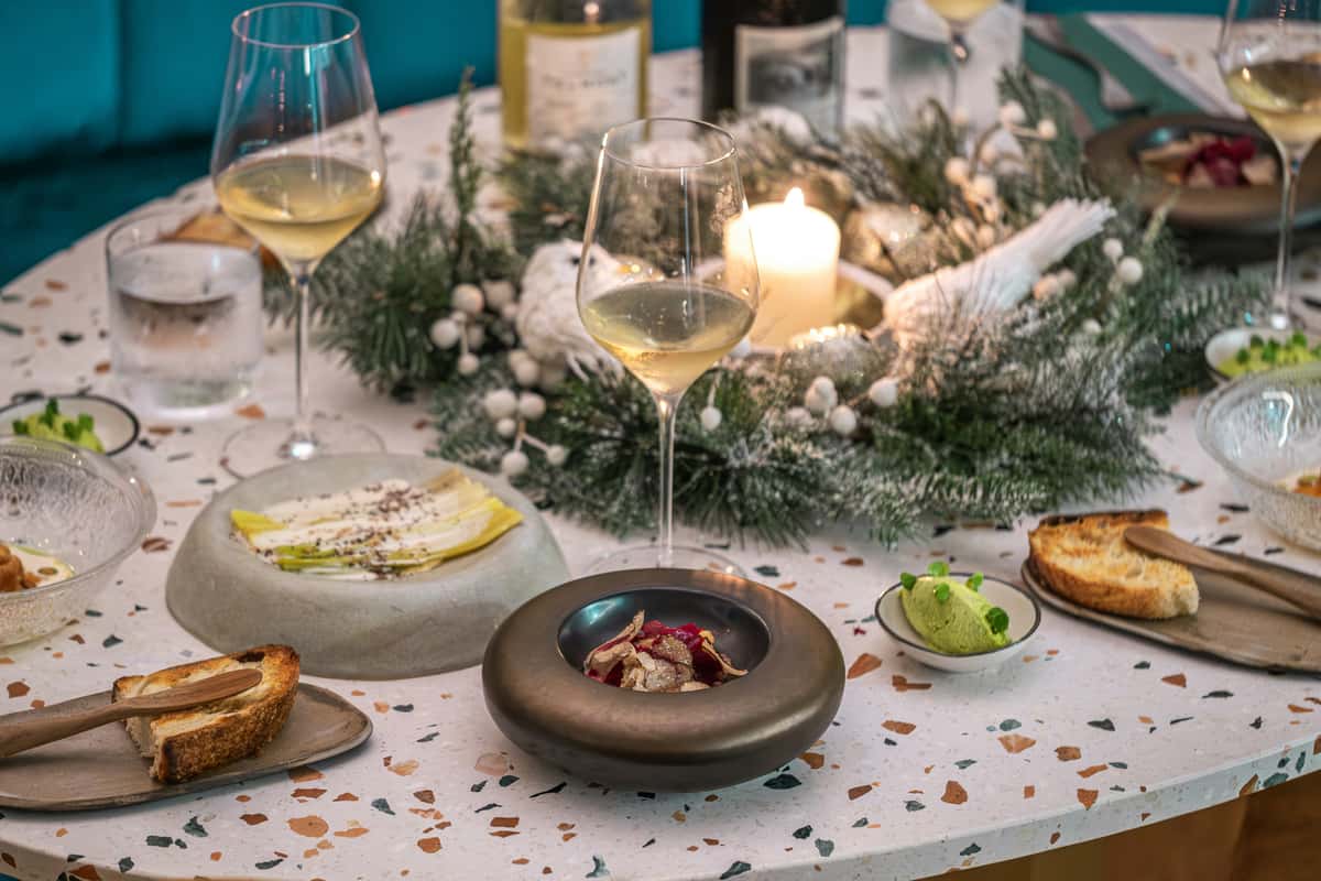 Elegant holiday table setting with white wine, gourmet appetizers, toasted bread, and a festive candlelit pine wreath centerpiece.