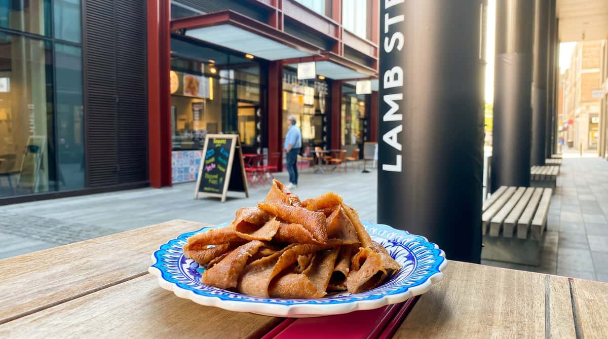 Shaved doner kebab meat on a blue and white decorative plate, set on a wooden table, with "LAMB STREET" sign and buildings in the background.