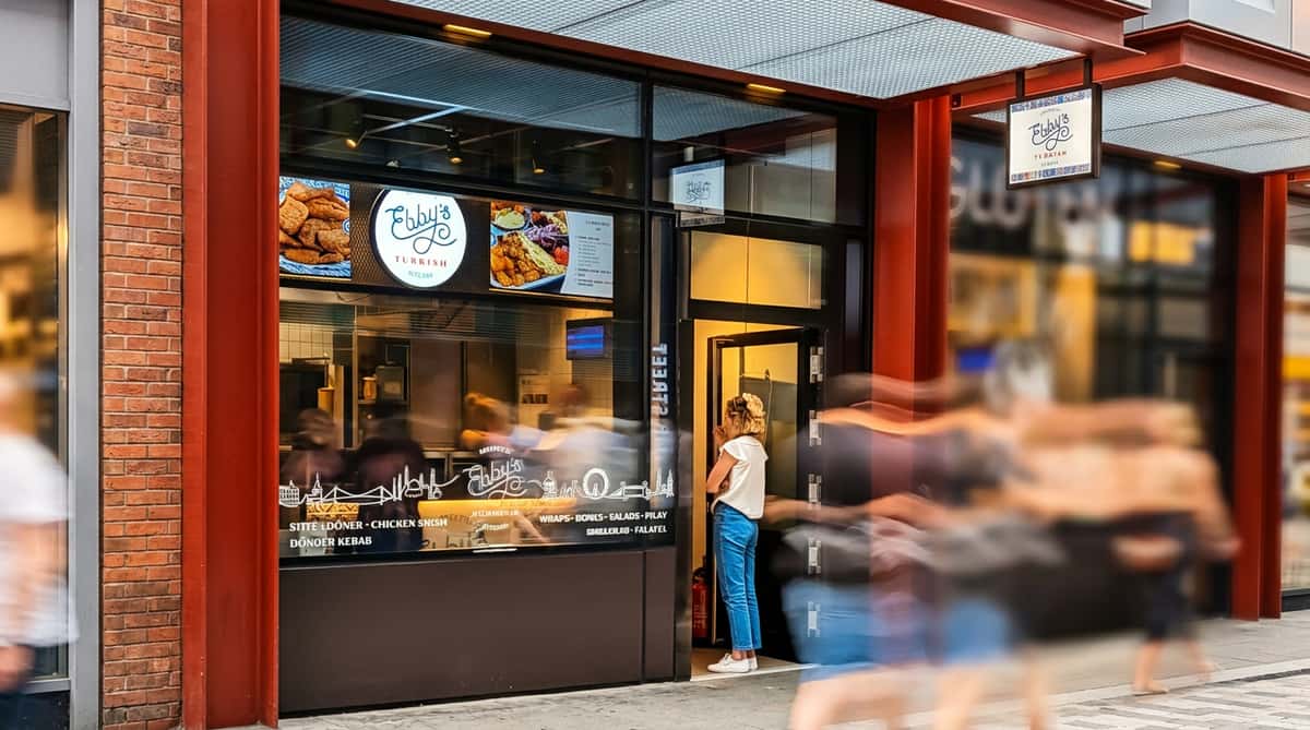 Storefront of Ebby's Turkish Kitchen with food menu displays and a woman standing in the doorway.