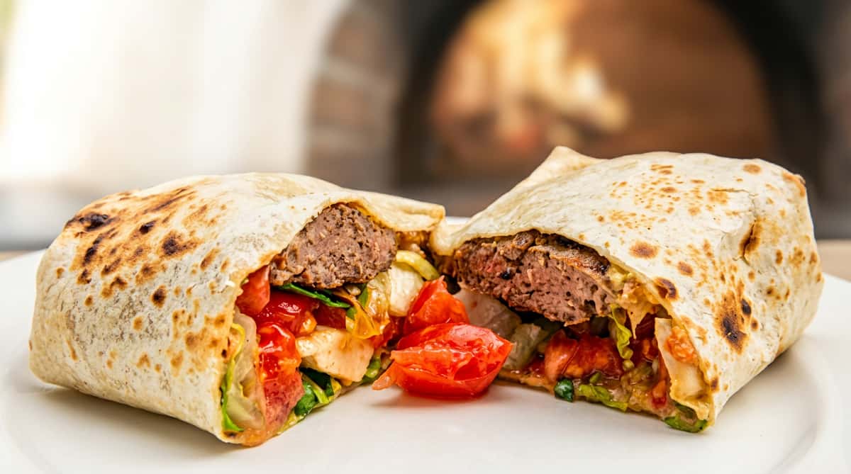 Grilled wrap, cut in half, revealing a burger patty, chopped tomatoes, and lettuce, in front of a fire oven.