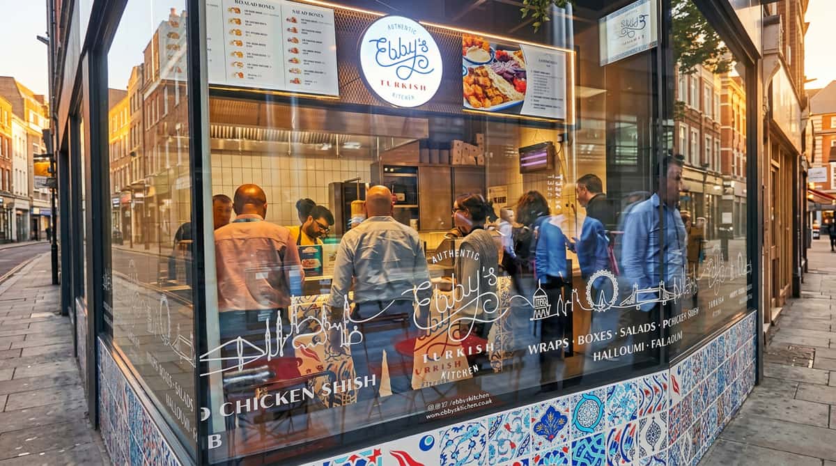 Exterior view of Ebby's Turkish Kitchen, showing customers and staff inside, reflected in a glass window with menu decals.