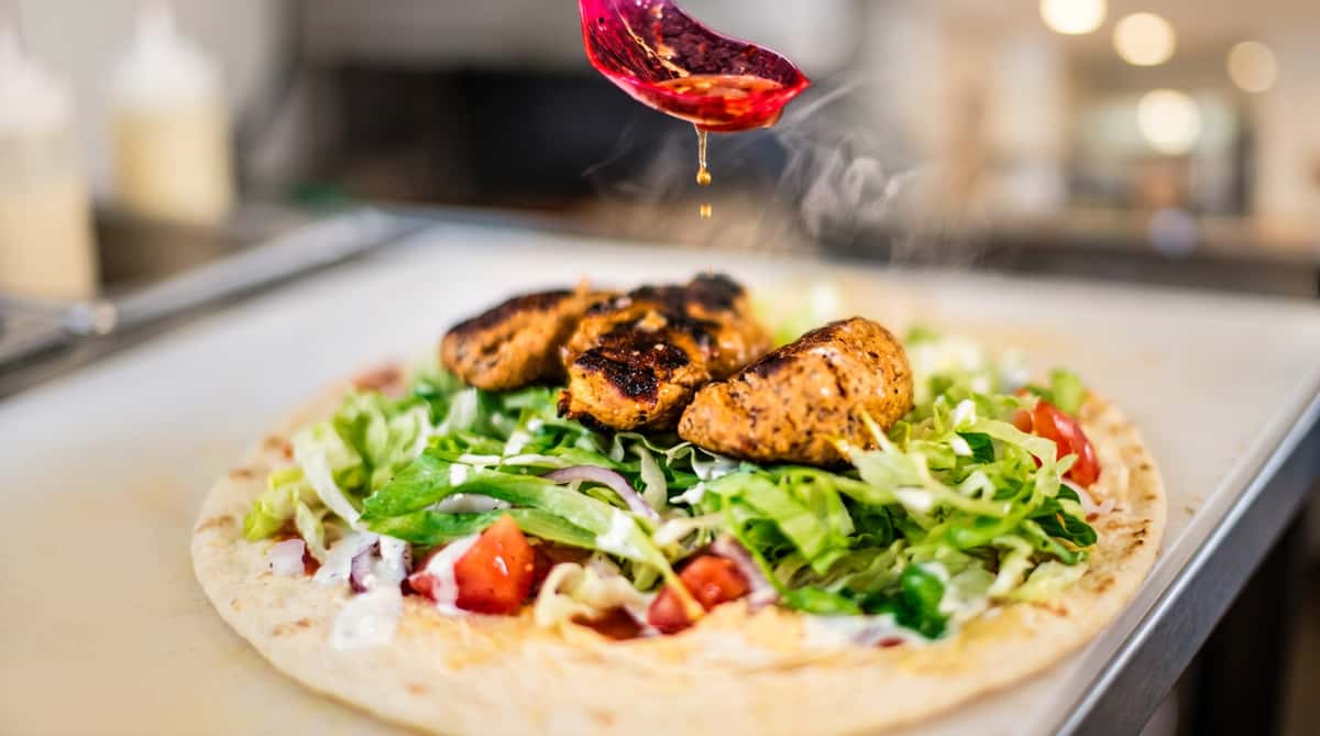 Kebab wrap with grilled meat, lettuce, tomato, red onion, white sauce, and dressing being drizzled from a red spoon.