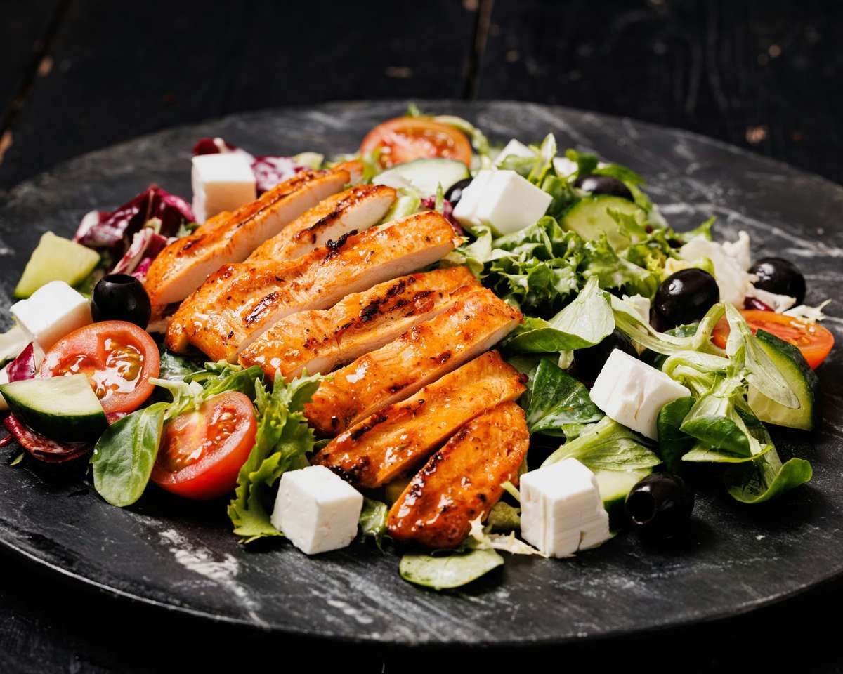 Grilled chicken breast slices on a mixed green salad with tomatoes, cucumbers, peppers, and creamy dressing on a dark plate