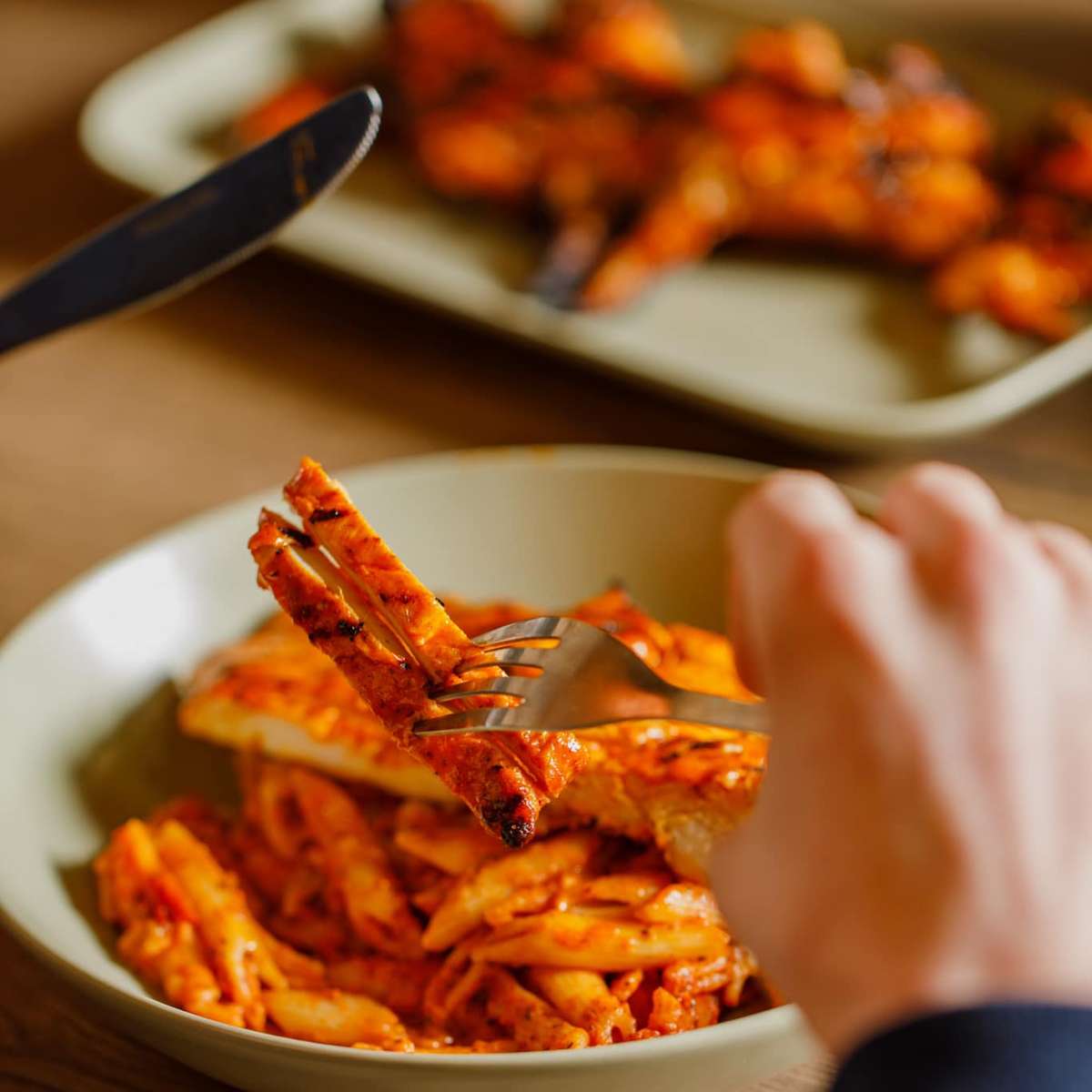 Fork lifting grilled chicken strips over a bowl of penne pasta in spicy tomato sauce, with plated chicken blurred in background