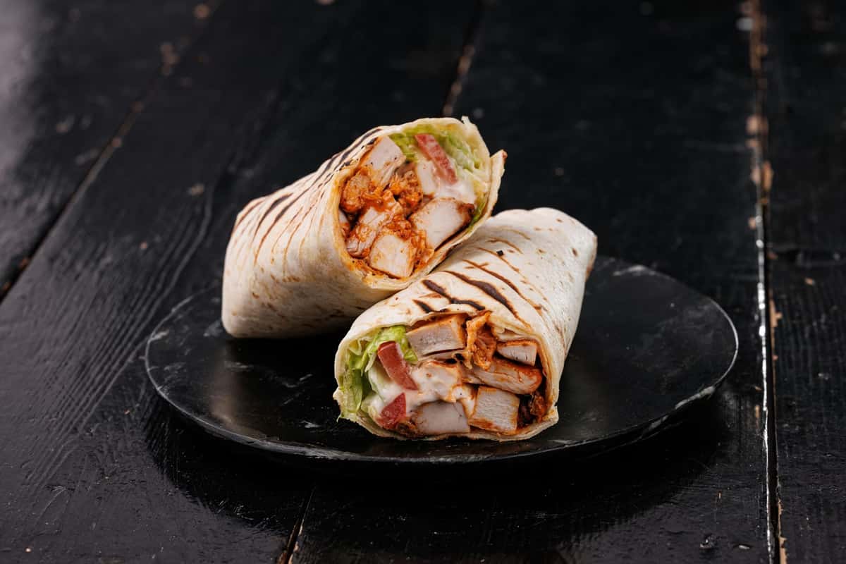 Grilled chicken wrap halves with lettuce, tomato, and creamy sauce on a dark plate against a rustic black wooden background