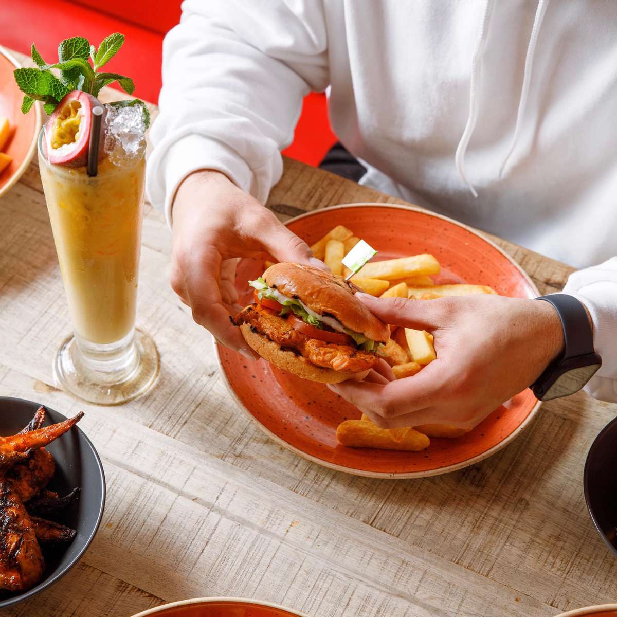 Hands holding a grilled chicken burger over a plate of fries on a wooden table, with a tall iced drink garnished with mint and passion fruit.