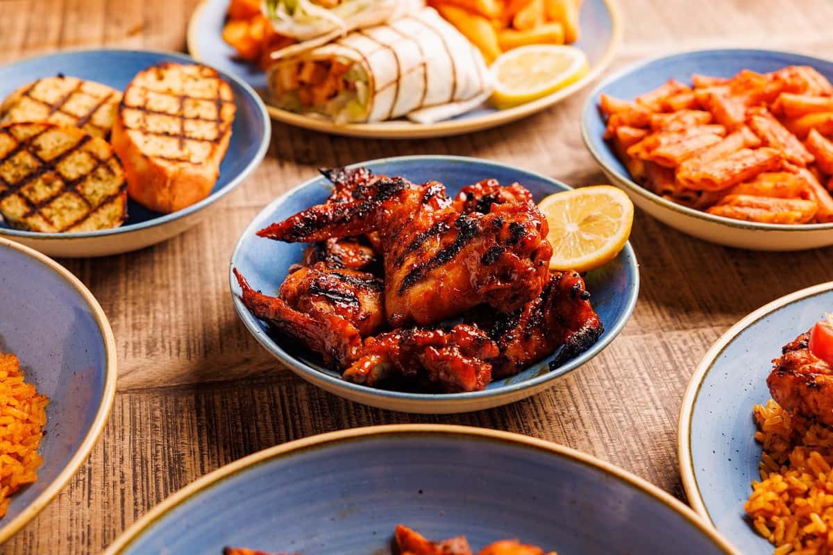 Assorted grilled chicken wings in bowls with lemon, fries, wraps, rice, and grilled vegetables on a wooden table.