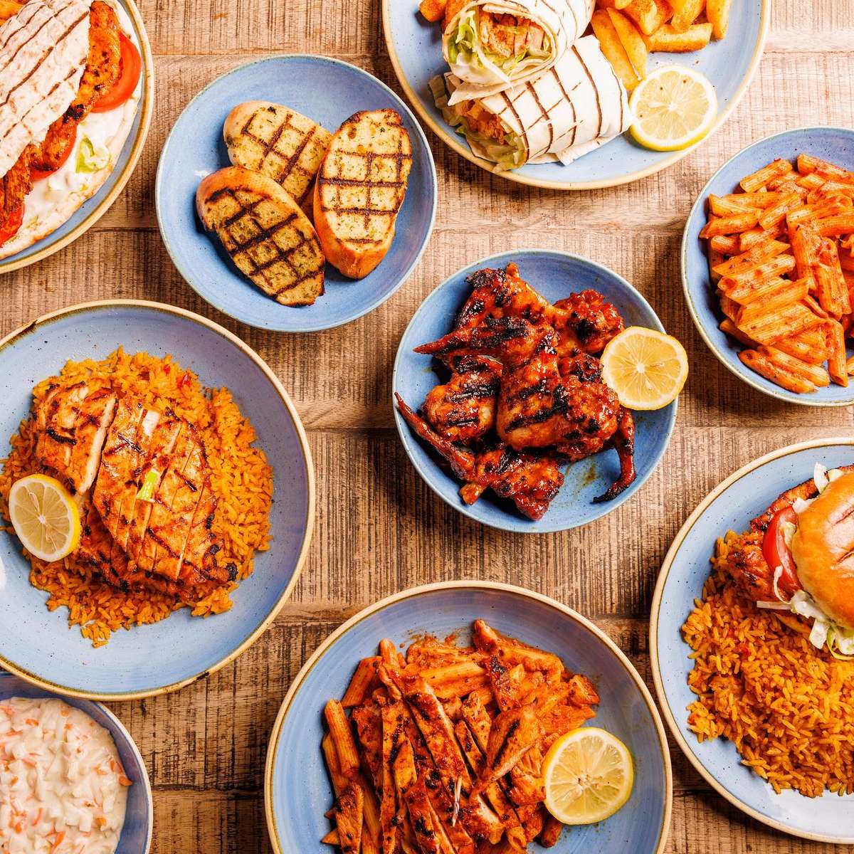 Overhead spread of grilled chicken, rice plates, pasta, wraps, burger, fries, and lemon wedges on blue dishes.