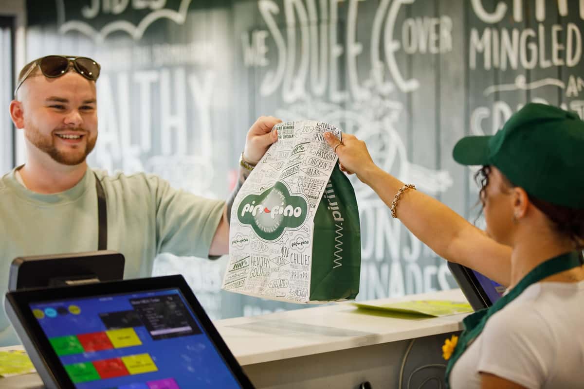 Smiling customer receives a branded takeaway bag from a staff member at a counter inside a casual restaurant setting