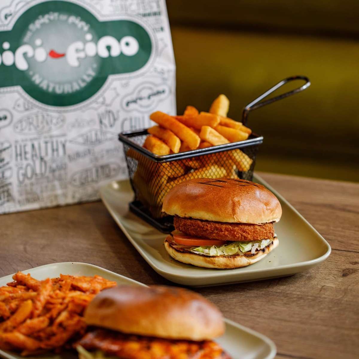 Breaded chicken burger with lettuce and tomato served with fries in a metal basket, with branded takeaway bag in the background
