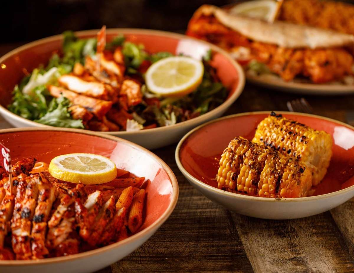 Assorted bowls of grilled chicken pasta, fresh salad with lemon, charred corn on the cob, and a chicken wrap on a rustic table