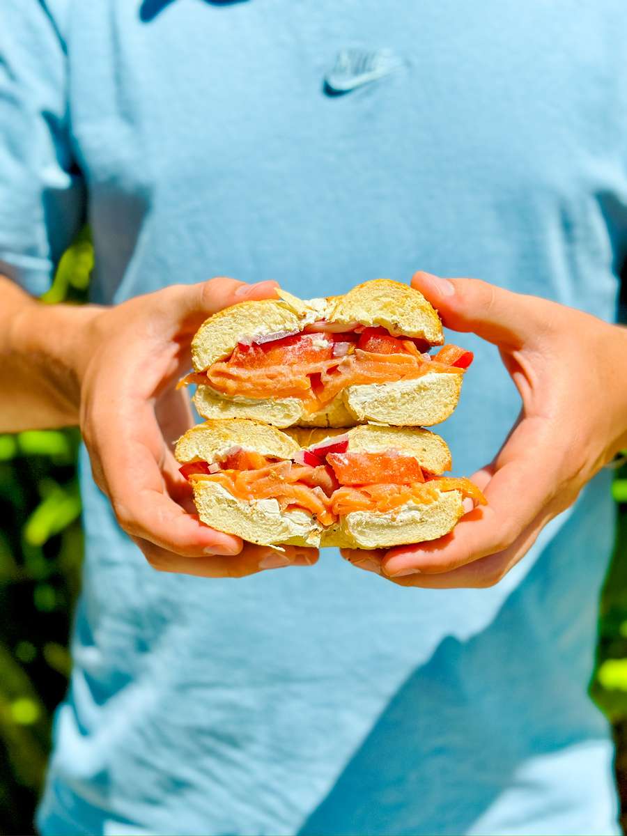 Close-up of hands holding a fresh lox and cream cheese bagel sandwich with tomato and red onion, shown against natural sunlight.