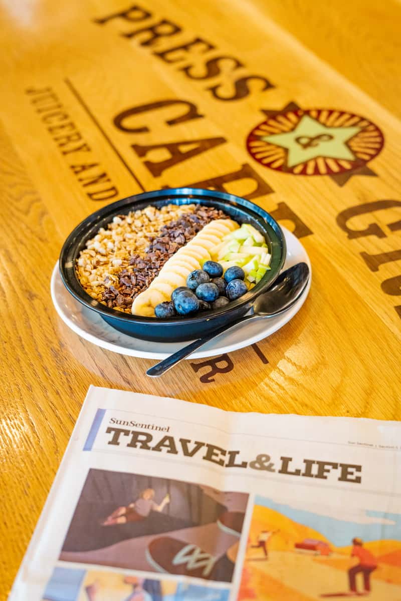 Colorful acai bowl topped with banana slices, granola, blueberries, and diced green apple, served on a wooden café table beside a travel newspaper.