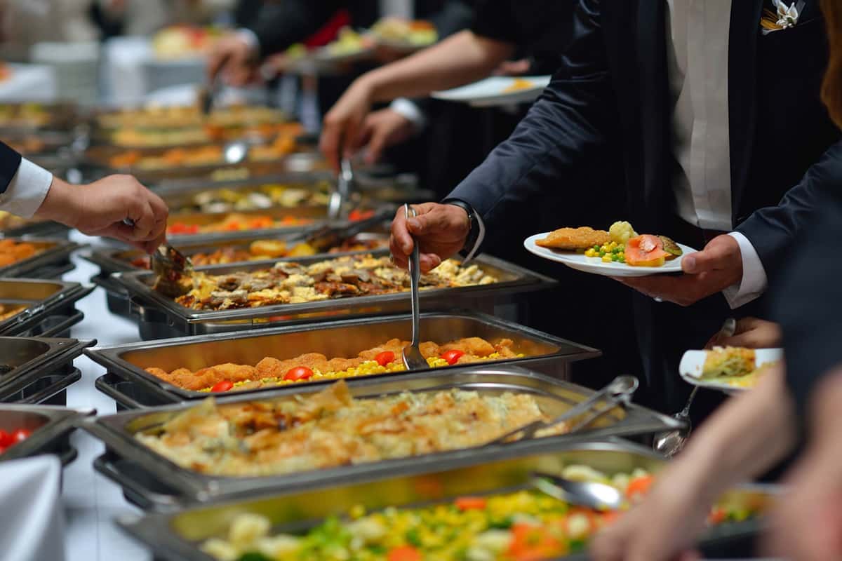 Guests in formal attire serving themselves from a buffet spread featuring a variety of hot dishes, vegetables, and breaded entrées.