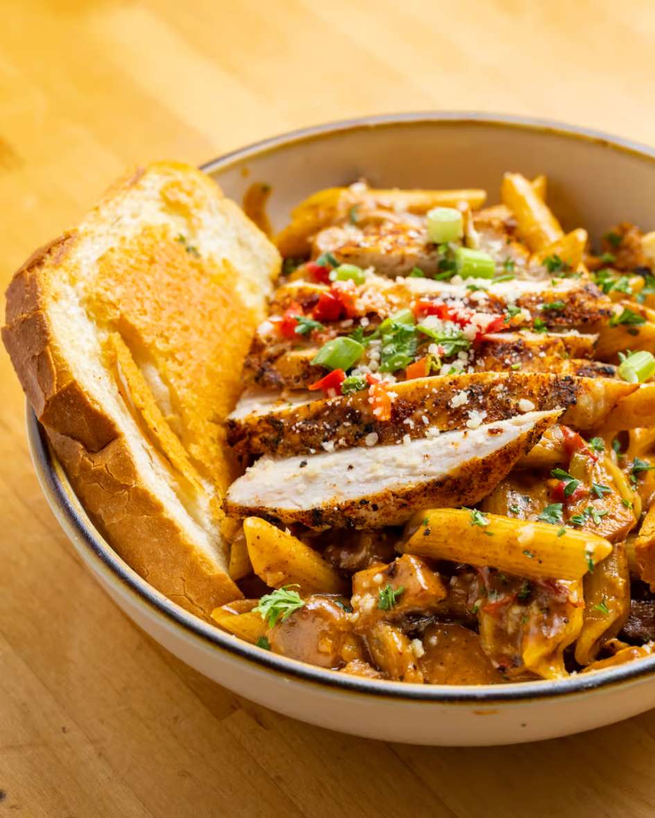 Creamy Cajun chicken pasta topped with sliced blackened chicken breast, garnished with diced bell peppers, scallions, and grated Parmesan, served with a thick slice of garlic Texas toast in a rustic bowl on a wooden table.
