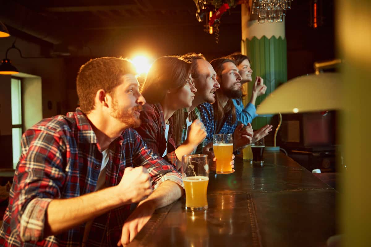 friends hanging out at the bar with glasses of beer in front of them, enthusiastically watching the tv