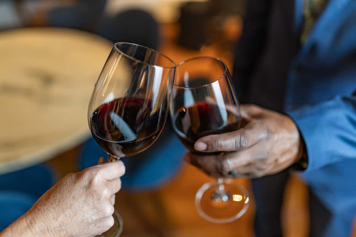 Close-up of two people clinking glasses of red wine in a toast, one person wearing a blue suit and the other with a bare arm, with a blurred background of a warmly lit indoor setting.