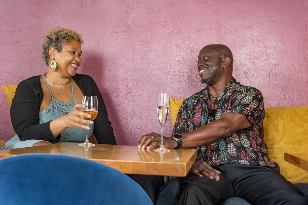 Smiling couple enjoying drinks at a cozy table with yellow cushions and a pink wall backdrop.