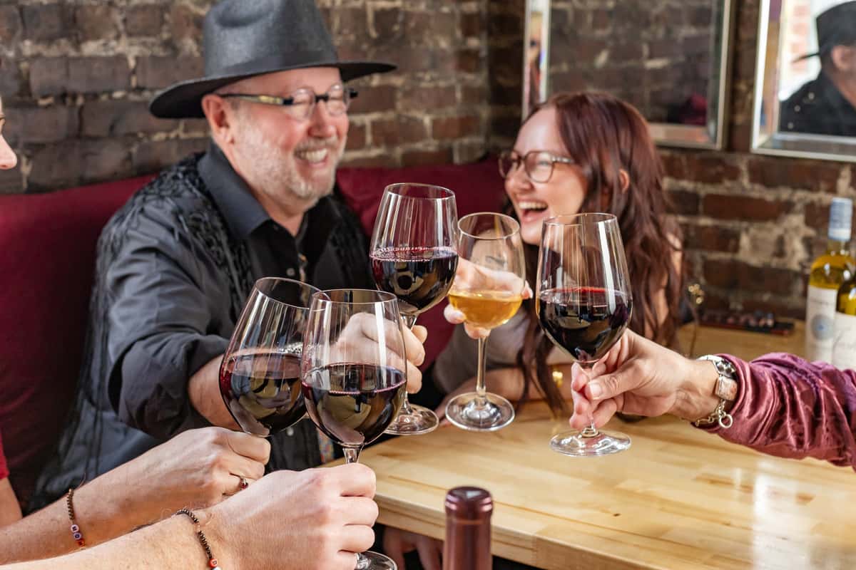 Group of friends laughing and clinking glasses of red and white wine at a cozy table with a brick wall backdrop.