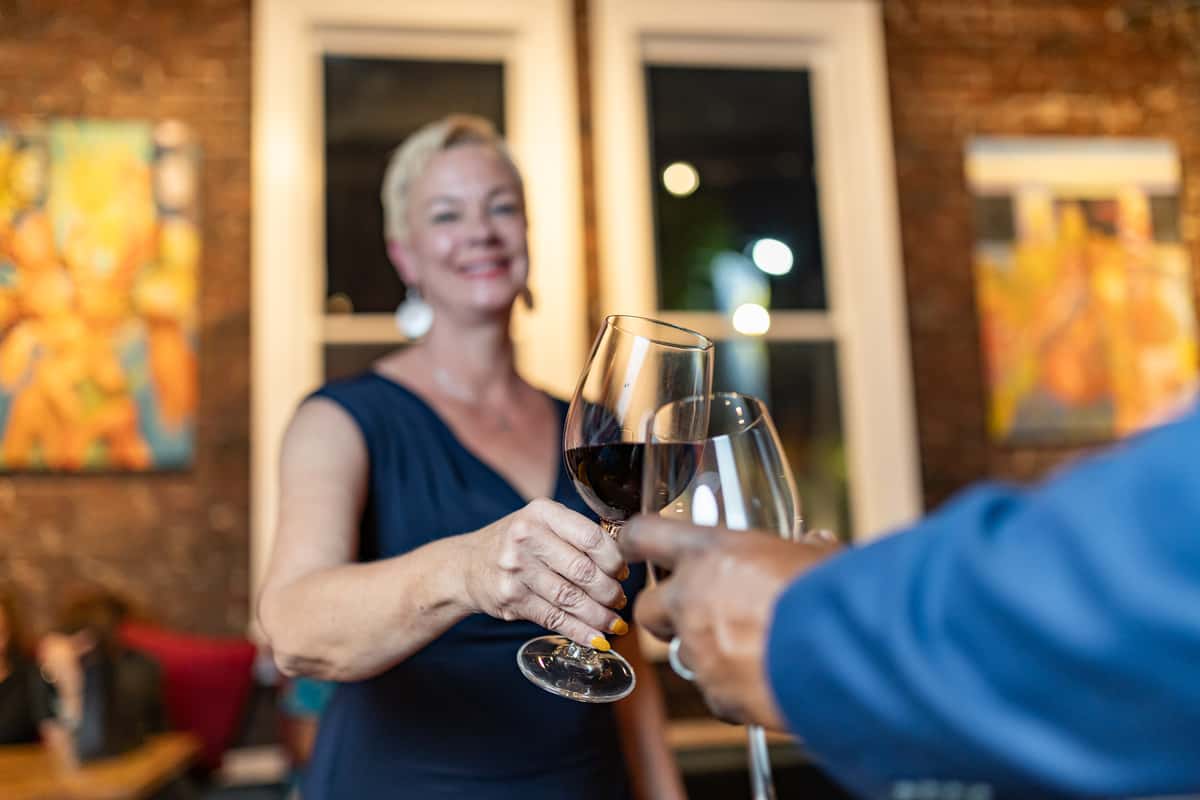 Woman in a navy dress clinks glasses of red wine with another person in a warmly lit, art-filled restaurant.