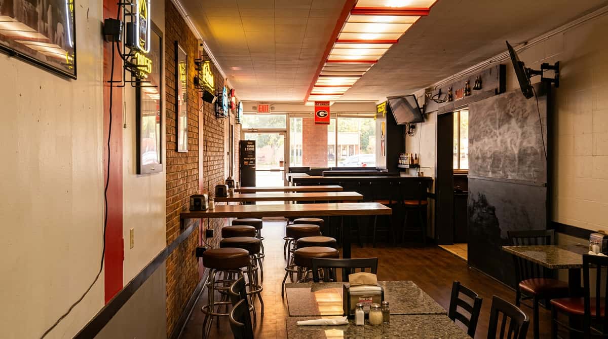 A narrow, dimly lit bar or restaurant interior with booth seating and tables, bright light streaming from the far end.