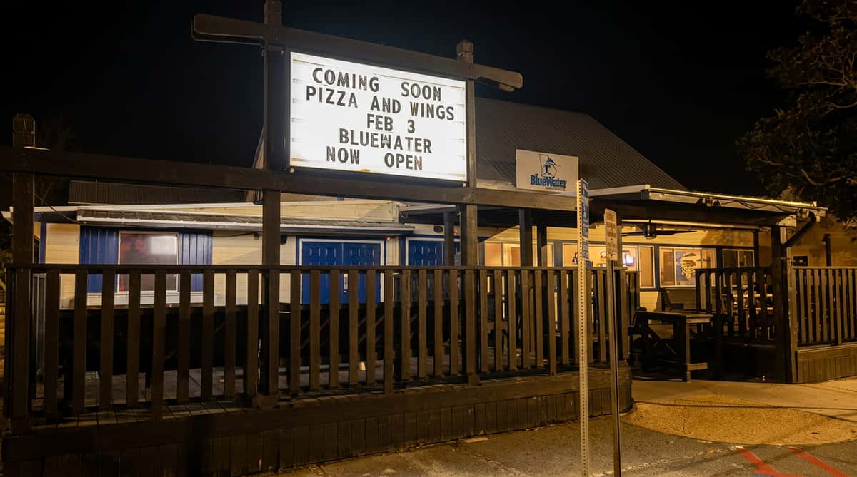 An illuminated sign at night announces "COMING SOON PIZZA AND WINGS FEB 3 BLUEWATER NOW OPEN" in front of a building.