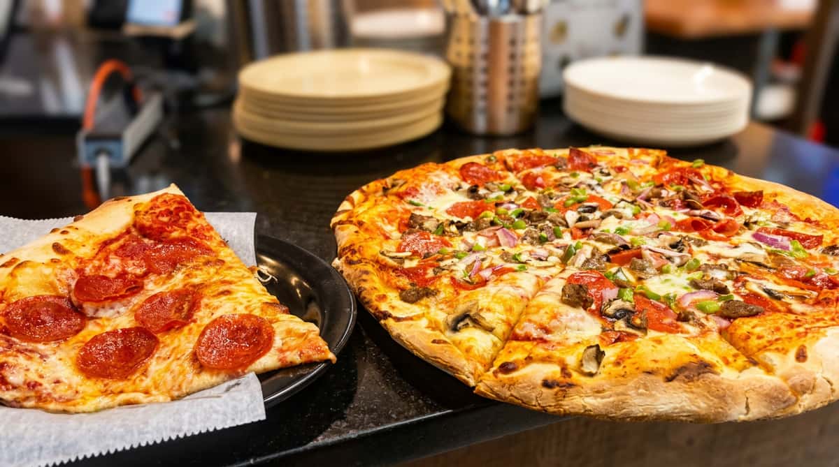 A whole pizza with pepperoni, mushrooms, green peppers, and red onions, next to a slice of pepperoni pizza.