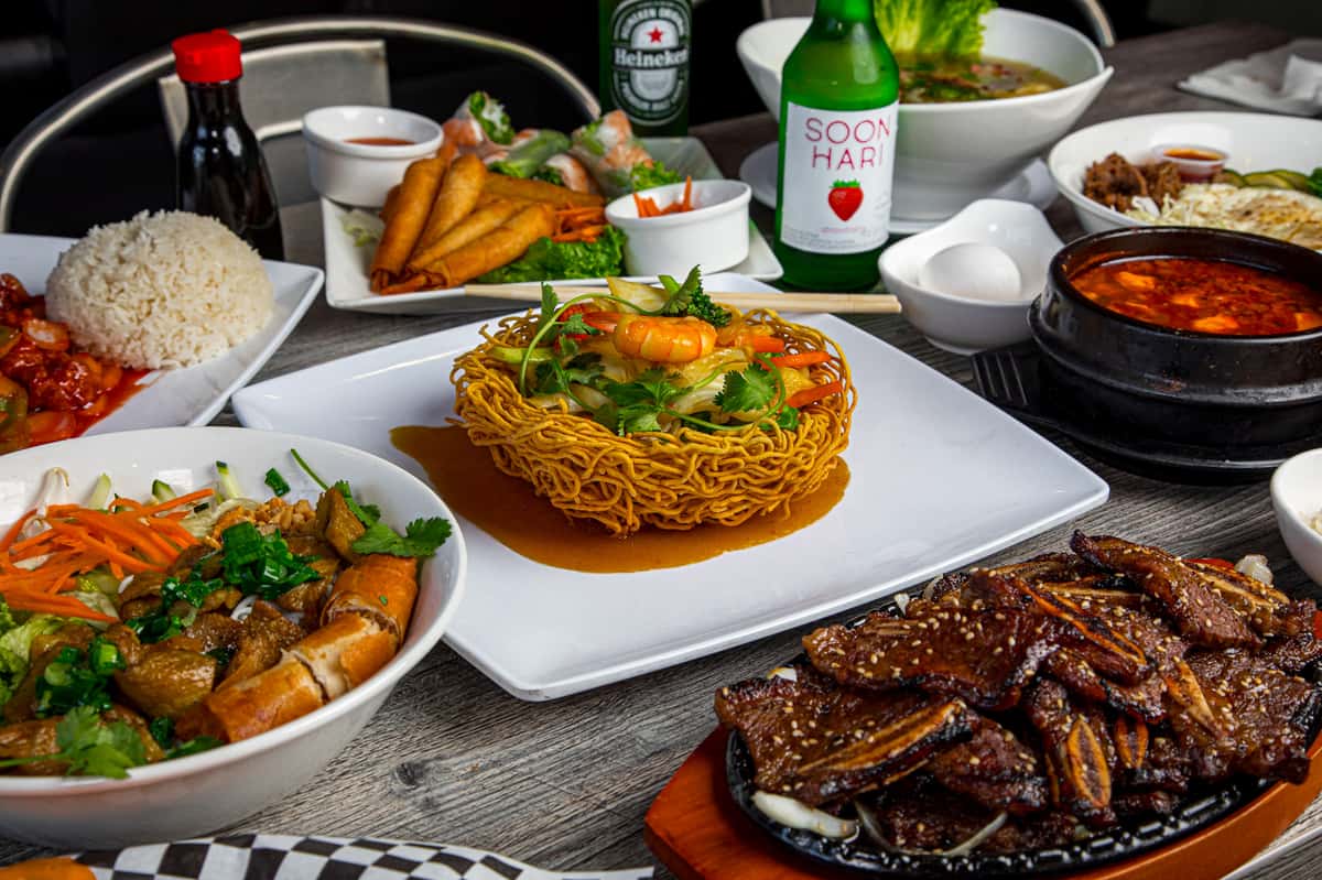 assortment of Vietnamese dishes featuring a crispy noodle basket filled with shrimp and vegetables, surrounded by spring rolls, grilled meats, and beverages, perfect for sharing.