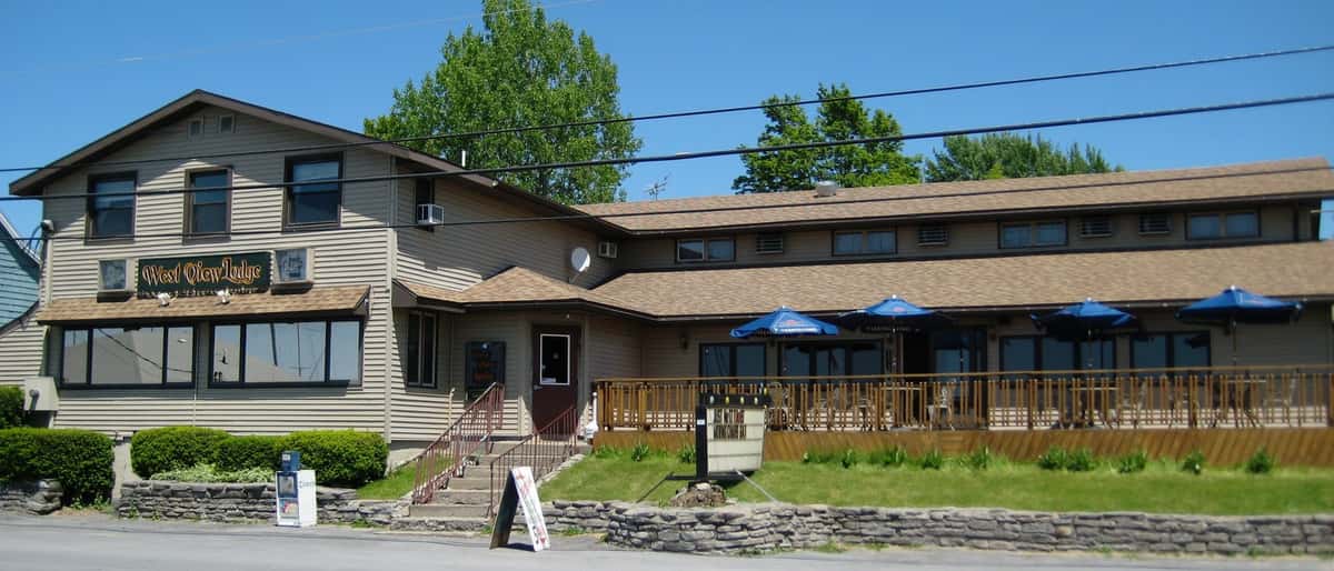 Exterior of West View Lodge, a rustic-style restaurant and bar with outdoor seating under blue umbrellas, located along a quiet roadside with lush greenery and a casual, welcoming atmosphere.
