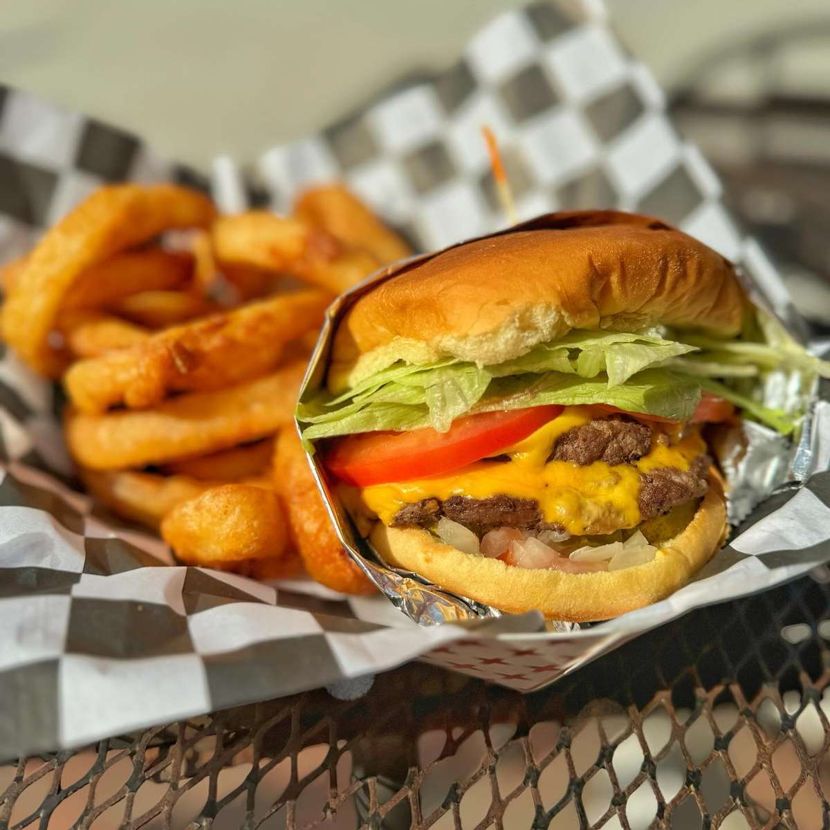 Cheeseburger with lettuce, tomato, and onions served in a basket with crispy onion rings on checkered paper.