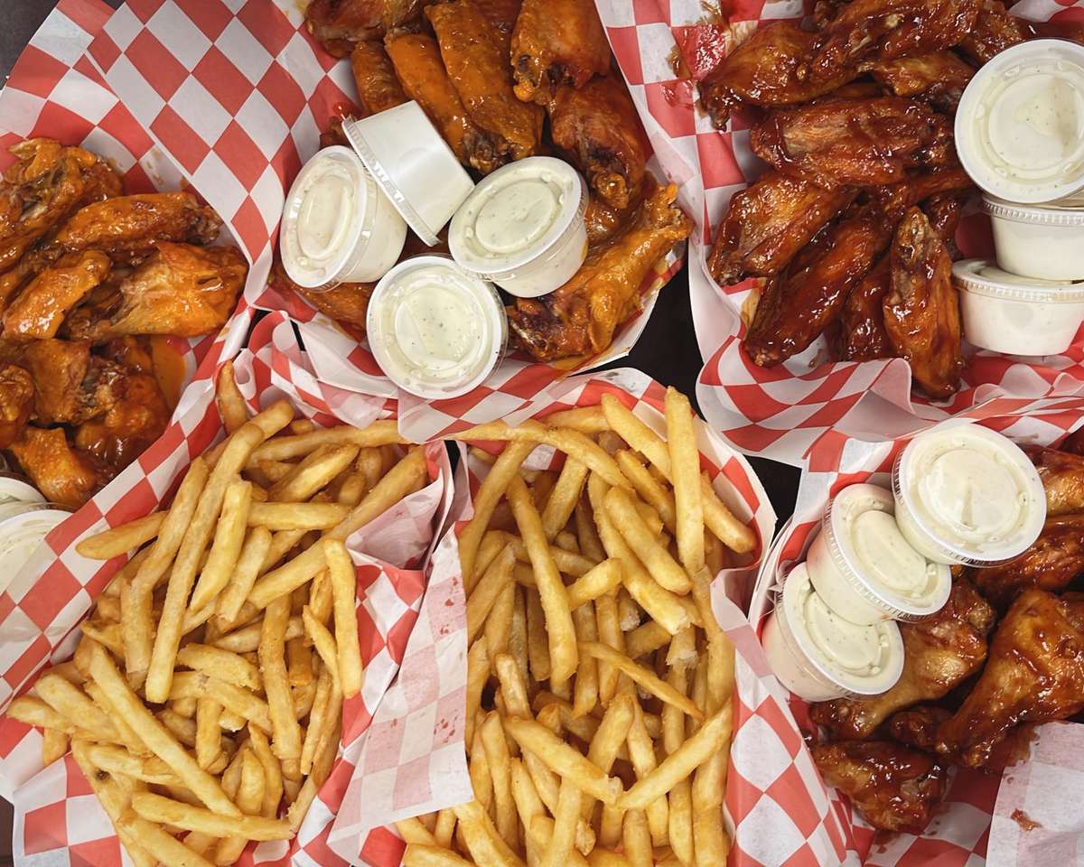 Assorted baskets of buffalo wings, BBQ wings, fries, and ranch dipping sauce served on red checkered paper.