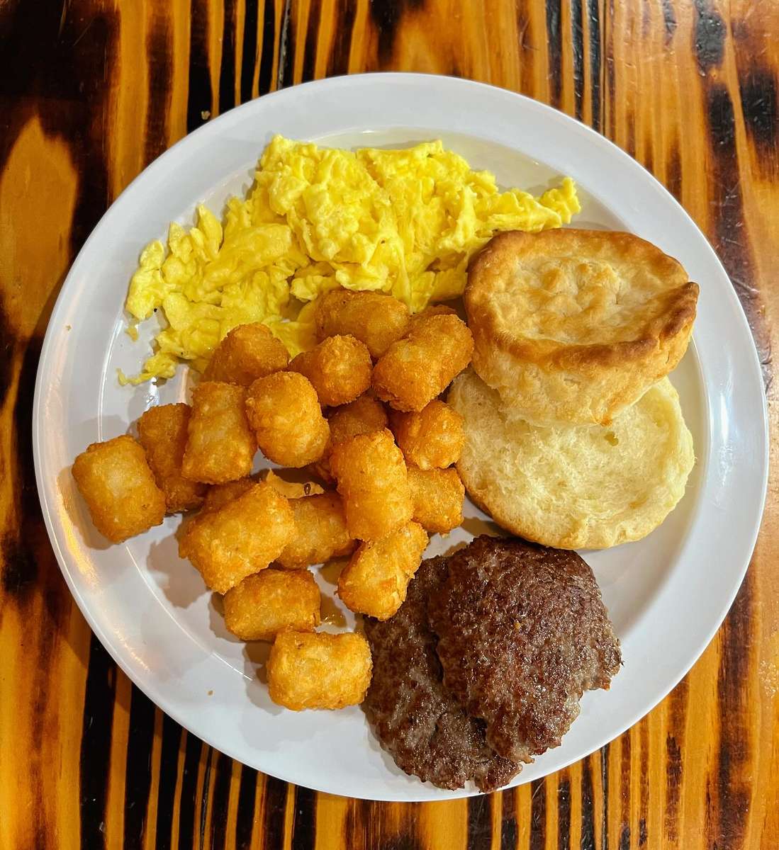 Breakfast plate with scrambled eggs, tater tots, sausage patties, and a split biscuit on a wooden table.