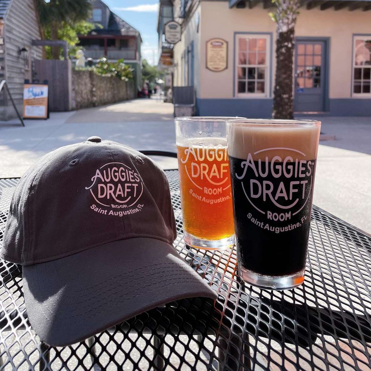 Two branded Auggie's Draft Room pints—one amber, one dark—sit on an outdoor table alongside a matching gray baseball cap, with a quaint St. Augustine street and palm tree in the background.