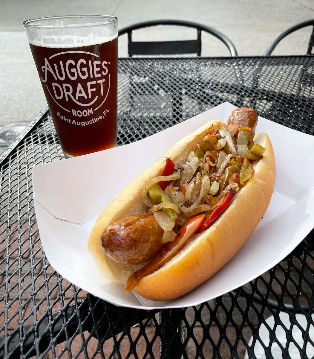 Grilled sausage topped with peppers and onions in a bun, served with a dark draft beer at an outdoor table.