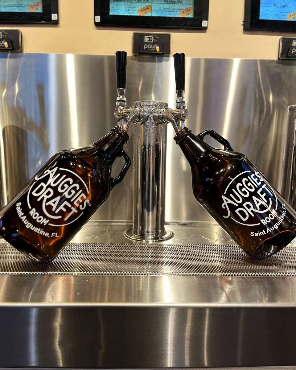 A pair of branded Auggie's Draft Room growlers are propped on a sleek stainless steel beer tap station, ready to be filled with fresh draft brews.