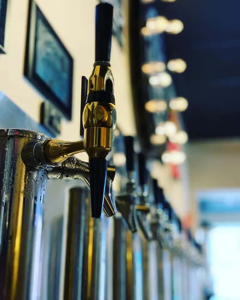 Close-up of a gold beer tap handle at a self-pour draft wall, with soft lights and other taps blurred in the background.