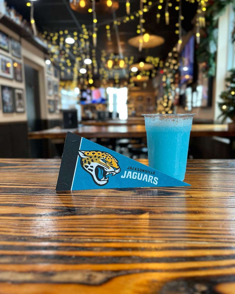 Blue frozen drink beside a Jacksonville Jaguars pennant on a wooden table in a festive sports bar decorated with string lights.