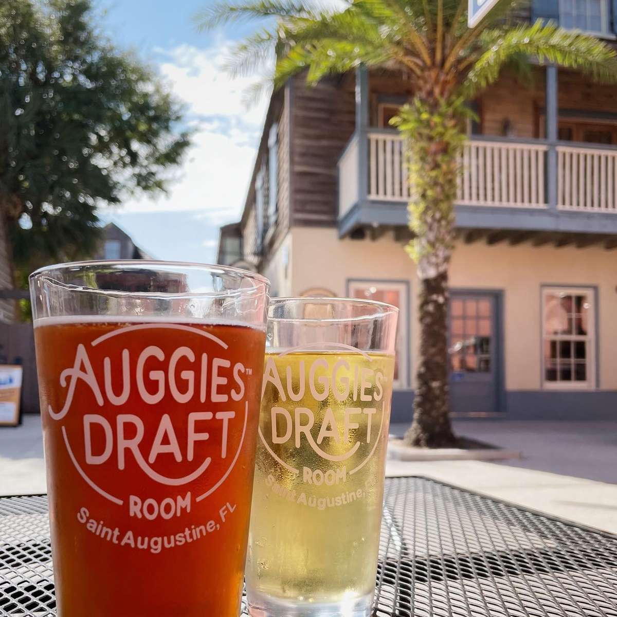 Two cold draft beers — one amber, one light — served in branded Auggie's Draft Room glasses, with a scenic backdrop of the bar's exterior and a tall palm tree in St. Augustine, FL.
