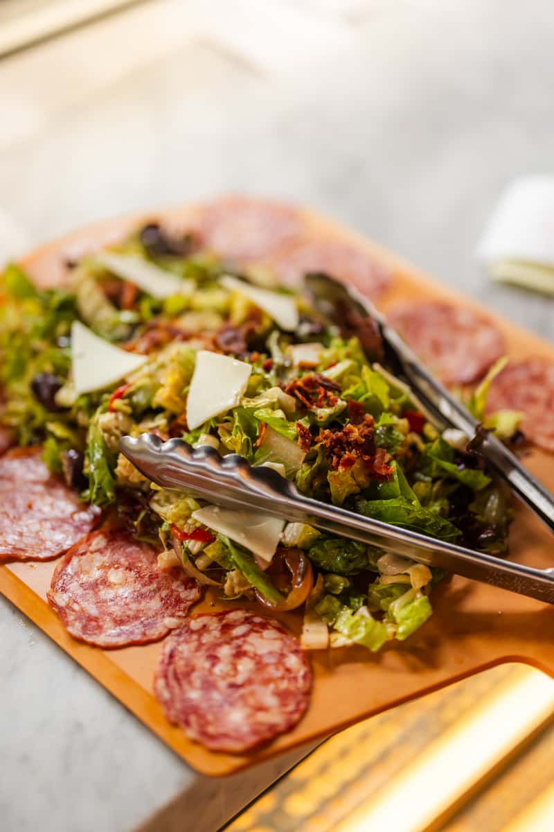 Fresh chopped Italian salad with shaved parmesan, sun-dried tomatoes, and mixed greens surrounded by slices of salami on a wooden board.