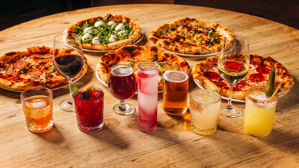 Assorted gourmet pizzas with a colorful lineup of craft cocktails, wine, and beer on a wooden table at a pizzeria