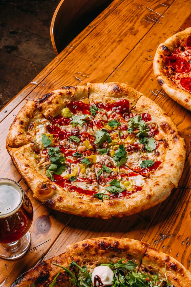 Wood-fired pizza topped with grilled chicken, banana peppers, fresh cilantro, and a drizzle of spicy red sauce, served on a rustic wooden table with a glass of amber craft beer.