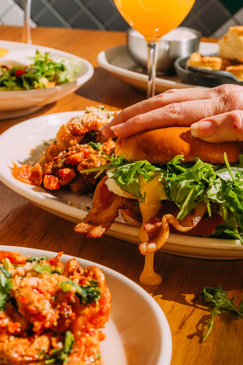 Close-up of a hand pressing down on a loaded breakfast sandwich with bacon, egg yolk dripping, arugula, and tomato, served with crispy potatoes on a brunch table