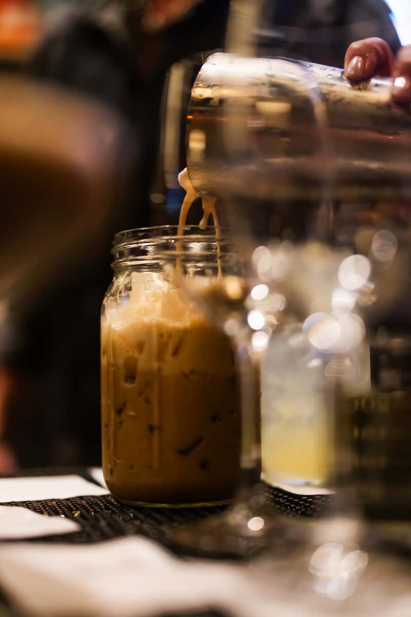 Bartender pouring an iced coffee cocktail into a mason jar at a dimly lit bar.