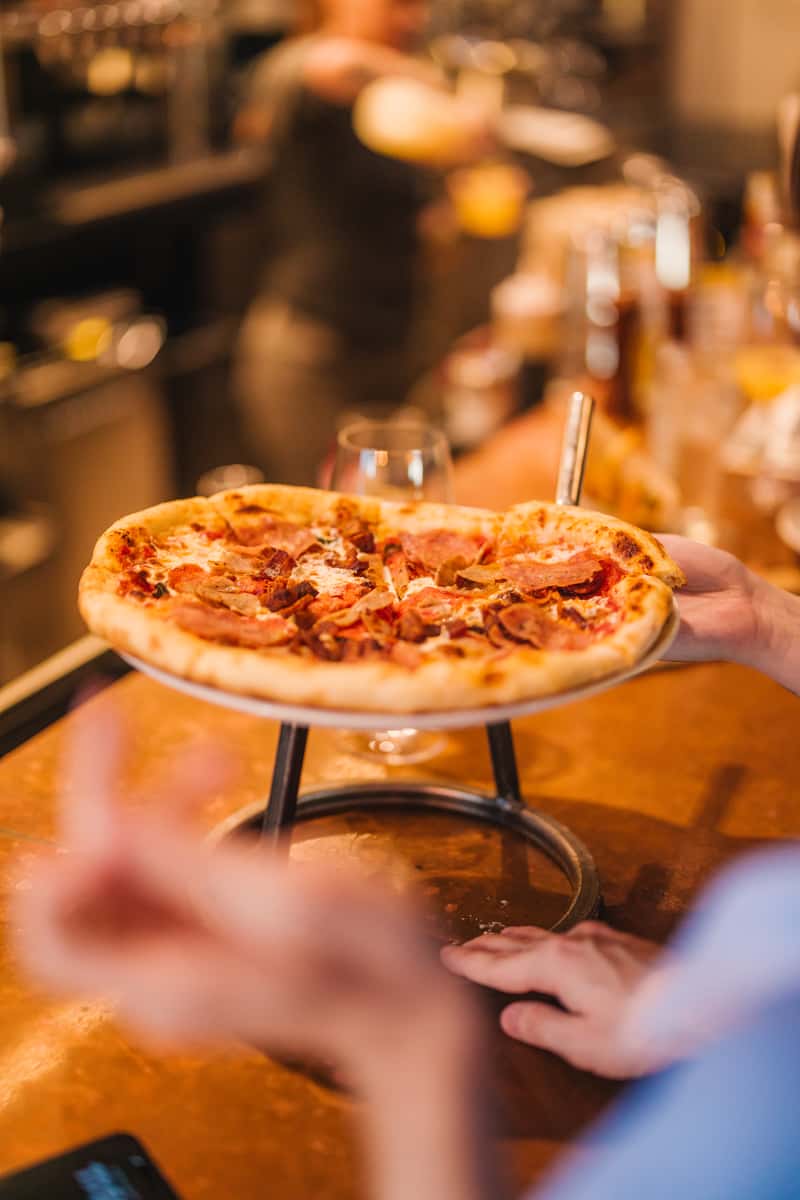 Freshly baked pepperoni and sausage pizza being served on a raised metal stand at the bar of a lively pizza restaurant.