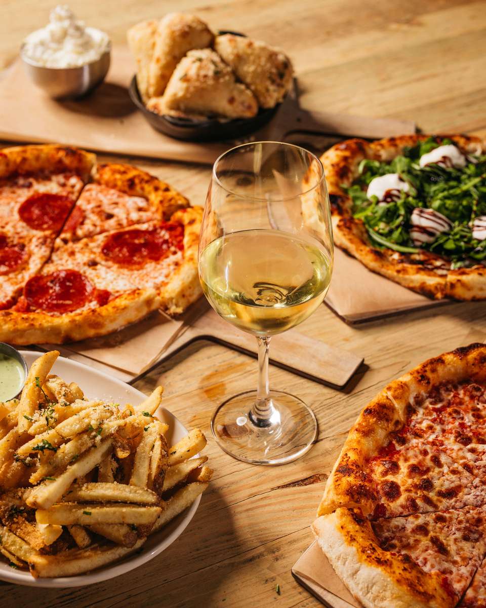 Table spread with gourmet pizzas, garlic parmesan fries, fluffy bread rolls with ricotta, and a glass of white wine on a rustic wooden table.